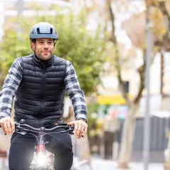 Mężczyzna w kamizelce i koszuli jedzie na rowerze, ma na głowie szary kask rowerowy.