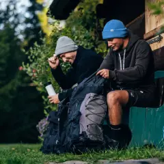 Dwóch przyjaciół z plecakami przygotowuje się do noclegu w lesie. Siedzą na ławce przed drewnianą chatką, otoczeni zielenią.