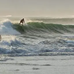 Surfer na desce ślizga się po zielonej fali, podczas gdy dwie sylwetki stoją w wodzie. To dowód, że surfing w Polsce jest możliwy!