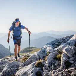 Mężczyzna w sportowym stroju z plecakiem i kijkami przemierza skalisty górski szlak. To przykład intensywnego **trekking vs hiking** w malowniczym krajobrazie.