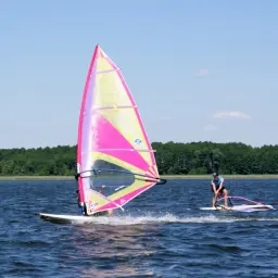 Dwóch surferów na jeziorze, jeden z kolorowym żaglem, cieszy się windsurfingiem na Mazurach.