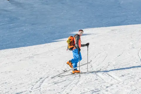 Mężczyzna w pomarańczowej kamizelce i niebieskich spodniach na nartach z fokami, gotowy na skitury. Co to jest? To przygoda!