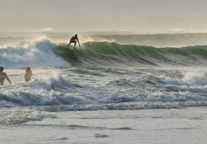 Surfer na desce ślizga się po zielonej fali, podczas gdy dwie sylwetki stoją w wodzie. To dowód, że surfing w Polsce jest możliwy!