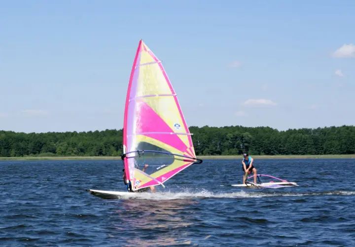 Dwóch surferów na jeziorze, jeden z kolorowym żaglem, cieszy się windsurfingiem na Mazurach.