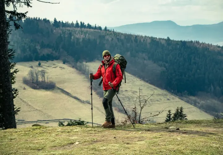 Mężczyzna w czerwonej kurtce i z plecakiem uprawia nordic walking na tle górskiego krajobrazu.