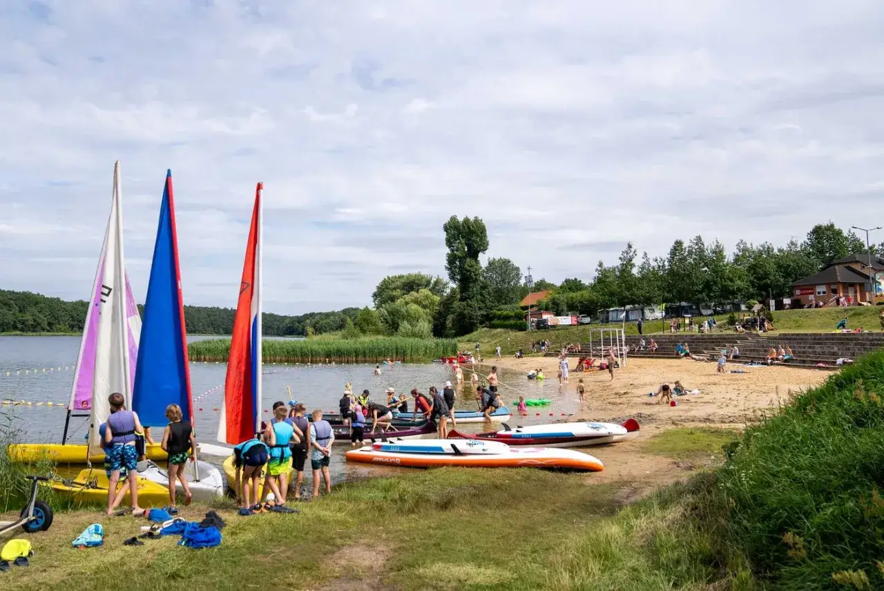 Dzieci i dorośli cieszą się aktywnym wypoczynkiem nad jeziorem. Żaglówki, kajaki i deski SUP czekają na wodzie i na plaży.