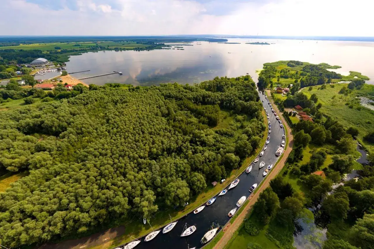 Malowniczy widok na Zalew Szczeciński z licznymi żaglówkami cumującymi wzdłuż kanału. Bujna zieleń otacza wodę, tworząc spokojny krajobraz.